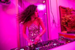 Samantha Michelle in a silver sequinned sleeveless dress, behind a DJ set-up at home, grooving away, with a pink filter to the image.