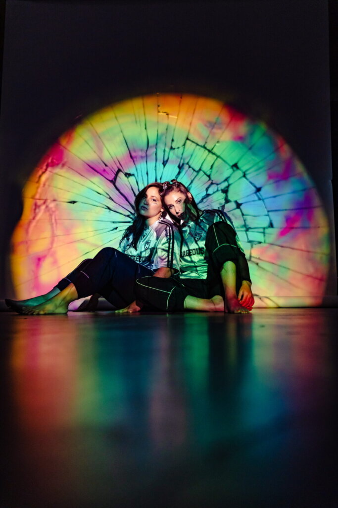 Royal She posing together on the floor with their backs to a wall as they are illuminated by a warped-colour spotlight.