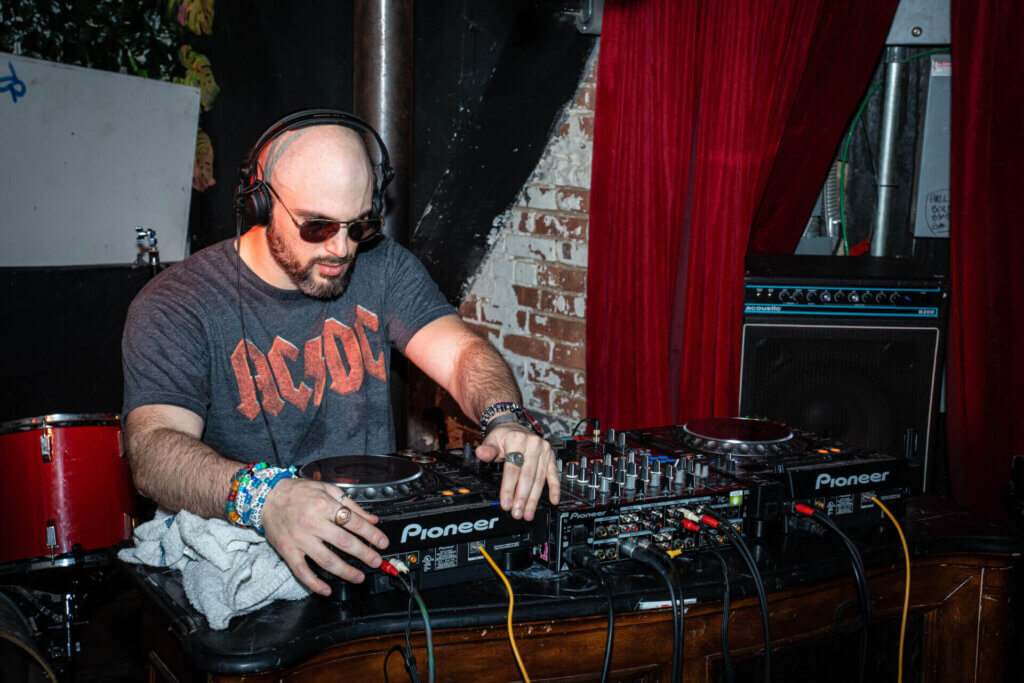 Mark Wise in a grey AC/DC t-shirt and dark shades standing behind a DJ deck, playing his music.