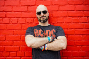 Mark Wise posing in front of a bright-red painted brick wall with his arms folded in front of him, wearing a grey AC/DC t-shirt and dark shades.