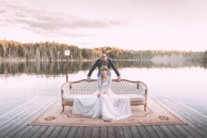 A gorgeous wintery-filtered photo of Somewhere in Between posing on a boardwalk on a lake with Deena Robertson wearing a beautiful wedding dress, sitting on a couch with Austin Bisnow standing behind her. A forest can be seen beyond the lake.