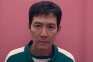 Screenshot from the trailer of Squid Game Season 2 which sees Lee Jung-jae reprising his role as Seong Gi-hun, as he re-enters the game, dressed in the green uniform and standing in front of a camera with a pink wall behind him.