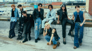 Stray Kids posing together outside in the daylight against a short white brick wall.