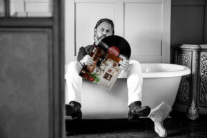 A black and white image of The Gleeman sitting in a bath with his legs over the side, holding a vinyl of his debut album with the cover of the LP being in full colour, bringing brown, red, and green colourisation.