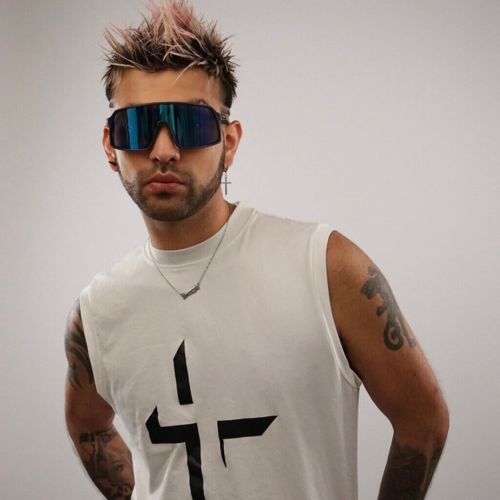 Jaime Adrian wearing a white tank top with a cross on it and a silver necklace and dark shades, posing for a professional photograph.