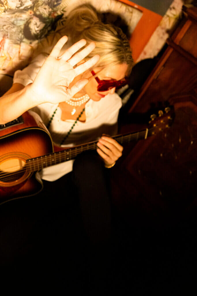 Bird sitting on the floor, turning her head away and holding up her hand to the camera, with her guitar across her lap.