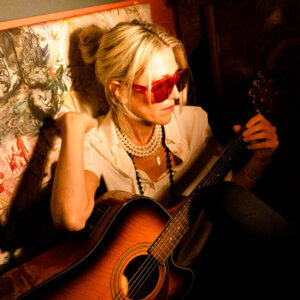 Bird, wearing a cream blouse, with her hair bunched up in a bun, along with a pearl-like necklace and red shades, holding a guitar across her lap with a Japanese-style art painting of trees and figures behind her.