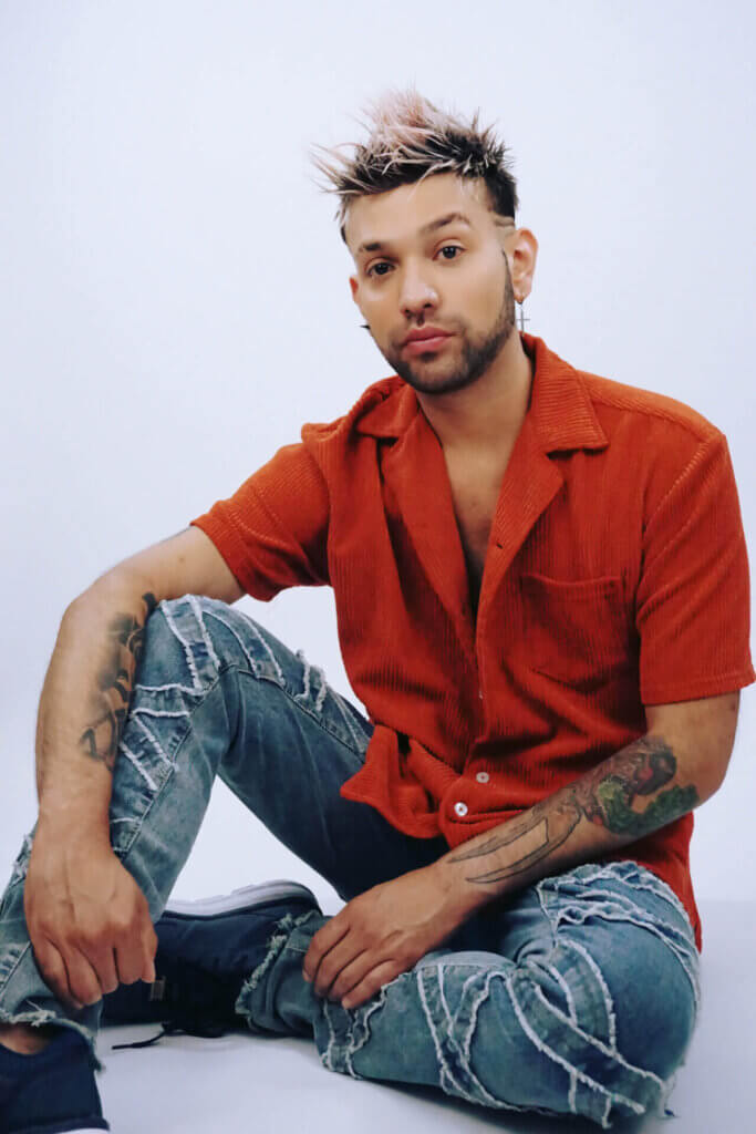 Jaime Adrian wearing an orange shirt with blue jeans and trainers, casually sitting on the floor, posing for a professional photo.