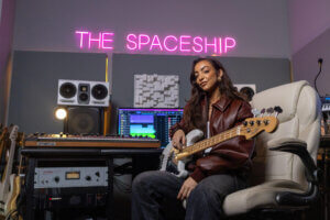 Promotional photo for Splice which sees Alissia sitting on a chair in a studio with a guitar on her lap, and there's pink neon lettering on the wall which says, "The Spaceship".