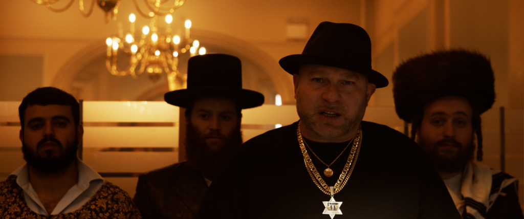 Still from the music video for "The Fathers Live" which sees four men cast in shadow with a candle chandelier behind them, as the guy in the forefront, who is Remedy, continues to rap.