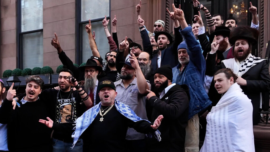 Still from the music video for "The Fathers Live" which sees Matisyahu, Remedy, and a group of their fellow Jews, pointing up to the sky, outside the synagogue, on the steps.