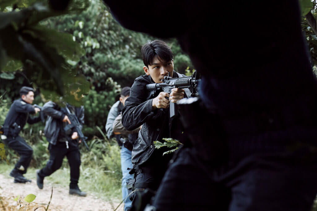 Still from Squid Game Season 2 which sees Wi Ha-joon playing Hwang Jun-ho, wearing black clothes and holding a gun in the middle of a wood land area.