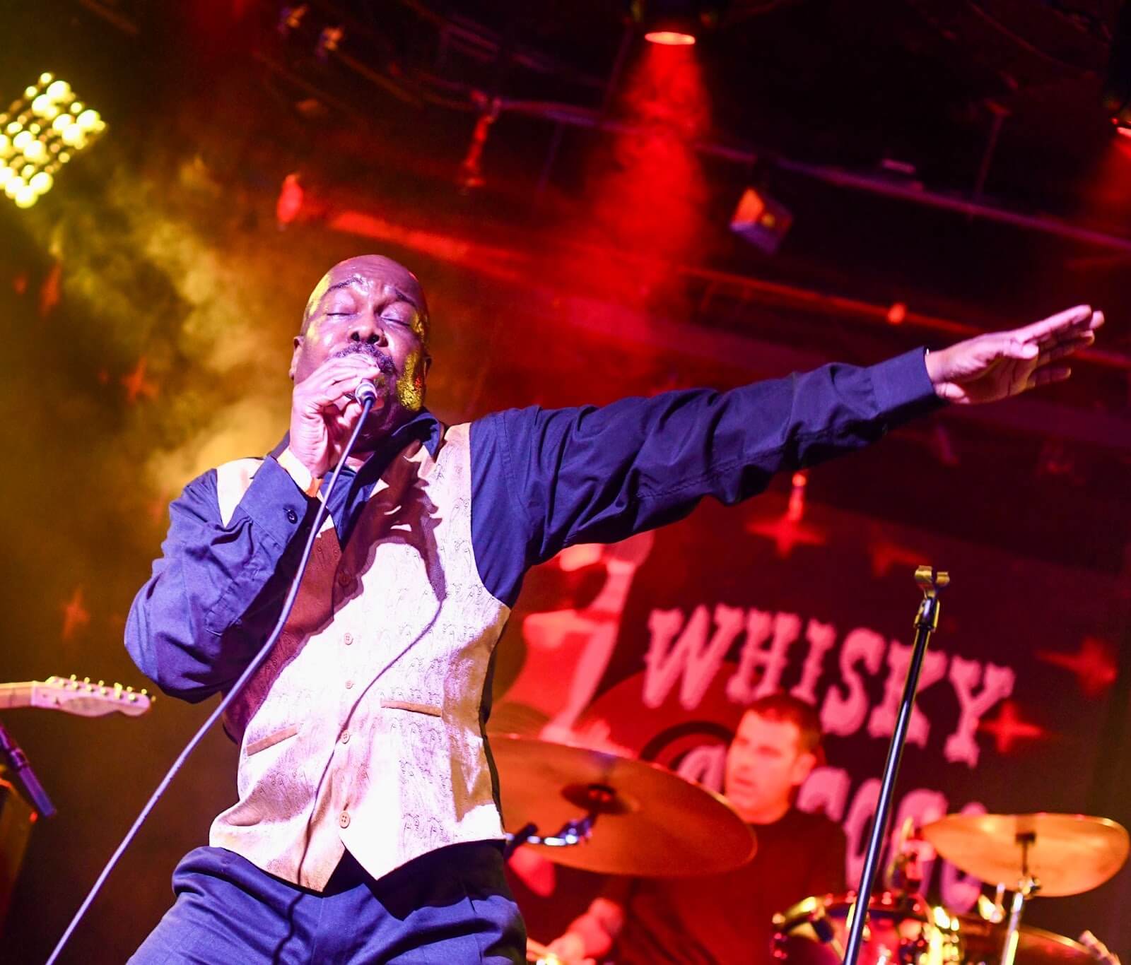 Kenneith Perrin singing into a microphone on stage wearing a white waistcoat over a blue shirt.