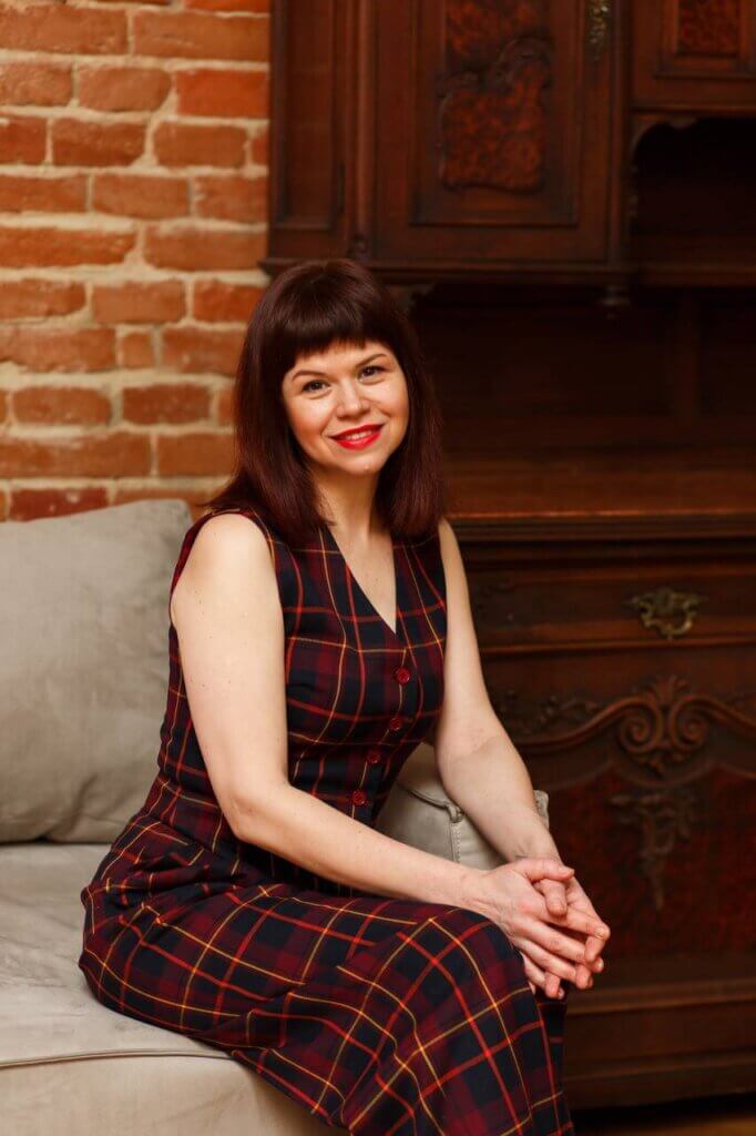 Liudmyla Kalabukha posing on a grey couch, wearing a dark almost-black but navy dress with red chequered lines on it.