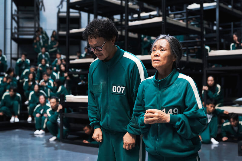 The mother and son duo from Squid Game Season 2 wearing the green tracksuit and standing in the home room area with beds and other contestants behind them.